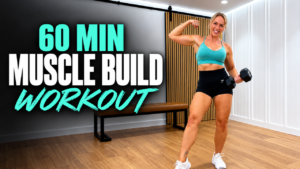 Woman over 40 flexing her bicep and holding a dumbbell beside a bench in a home gym, with the words 60 min muscle build workout, a full body dumbbell workout